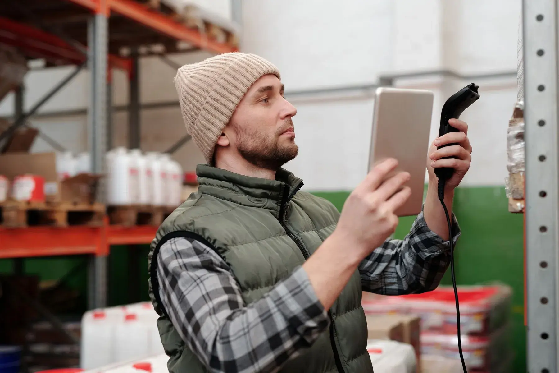 Warehouse worker in a beanie and vest scanning stock with a handheld scanner beside tall racking